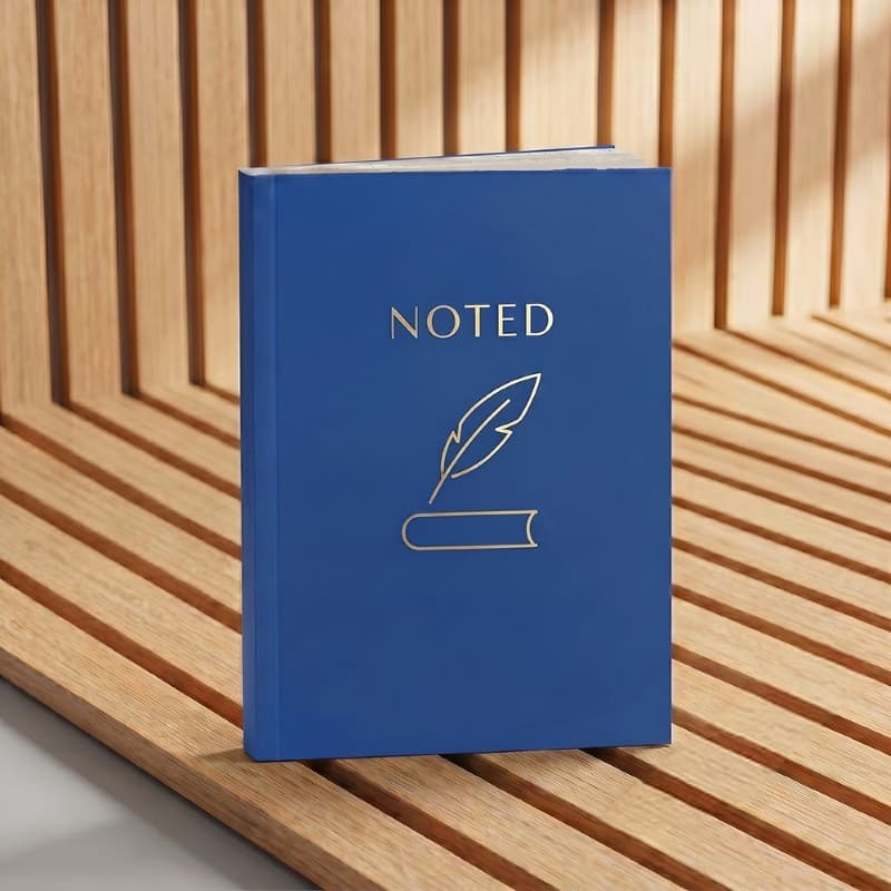 Blue soft bound notebook with "NOTED" and quill gold foil design, displayed on a modern slatted wood surface.