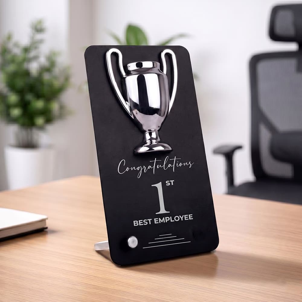 A black Winner Cup Desk Trophy with a chrome cup emblem and "1st Best Employee" inscription, standing upright on a corporate office desk.