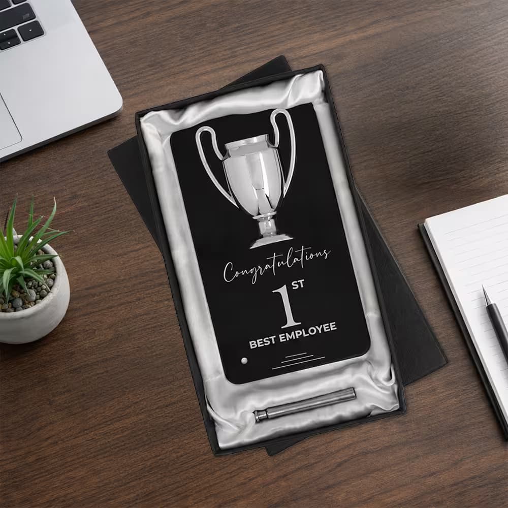A black Winner Cup Desk Trophy with a chrome cup emblem in a white satin-lined presentation box on a wooden desk with a laptop.