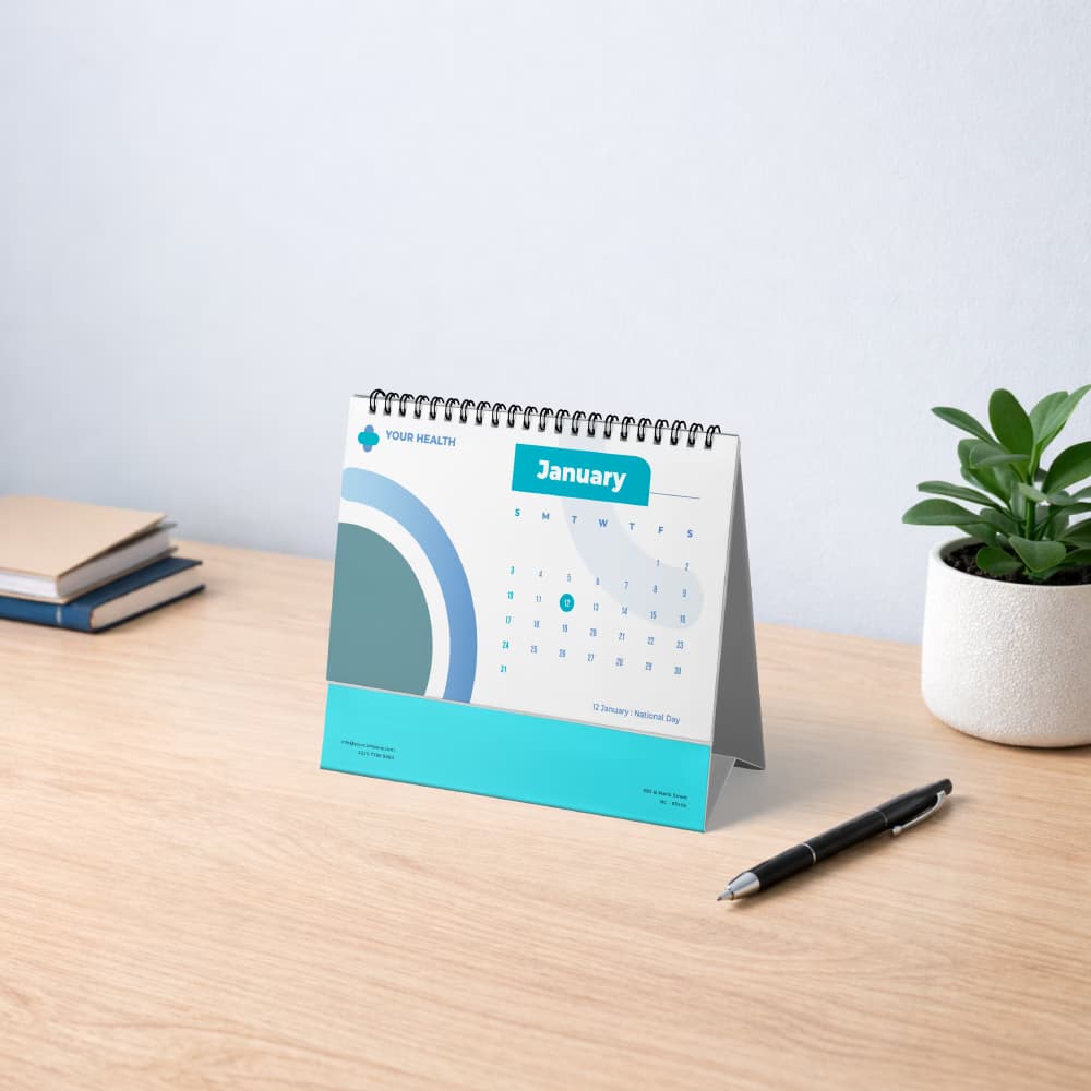 A landscape-oriented blue and white corporate desk calendar sitting on a bright office desk with a pen.